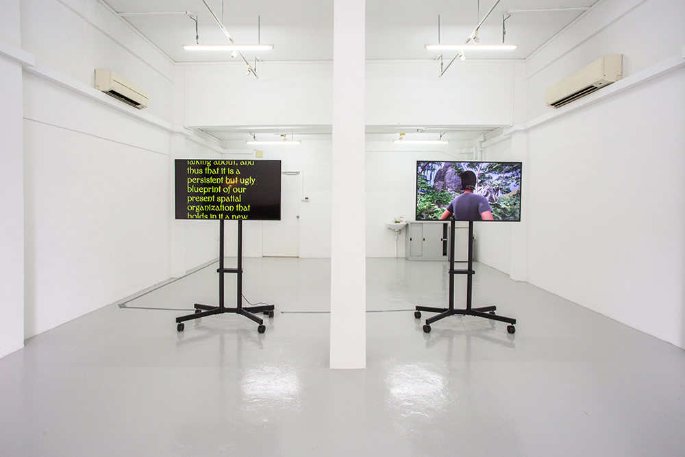 Exhibition view of two screens featuring text on the left screen and a female avatar on the right screen.