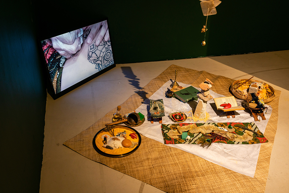 A screen showing henna being drawn on a body, miscellaneous items are on a woven mat in front of the screen.