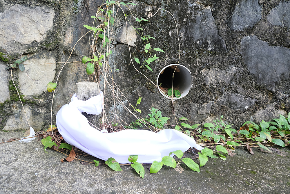 A white cloth is tied to a small concrete pillar next to a drain hole opening on the side of a concrete wall.