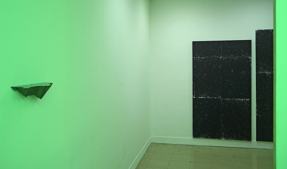 A room lit in neon in green, black canvas on one side of the wall and a sculpture on the other.