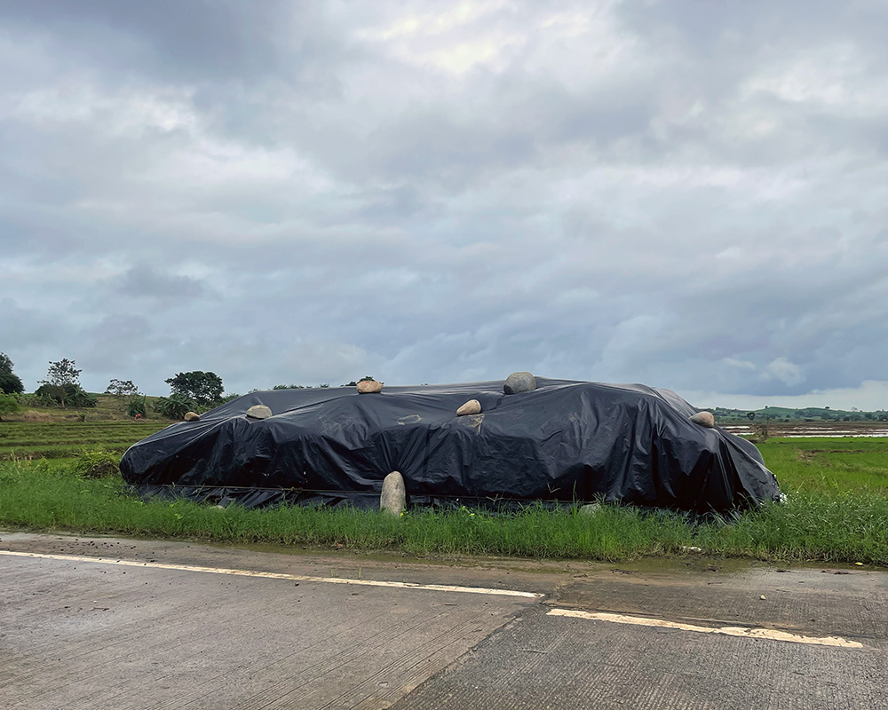 [Weatherproofing] A thick plastic sheet protects an unknown object or objects from the elements in an open field in the province of Isabela. The sheet is held in place by sizeable rocks.